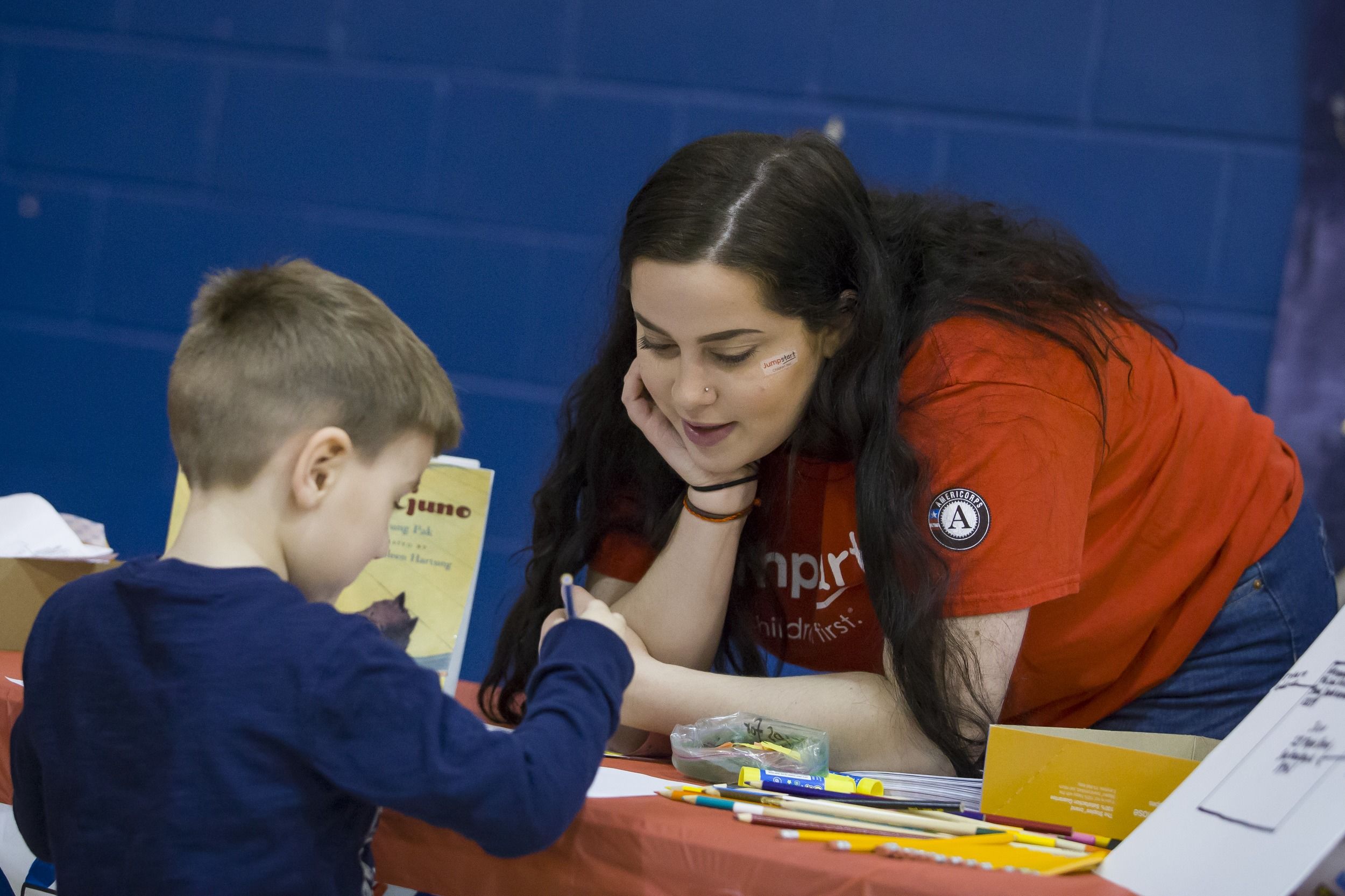 A student leans over a table to speak with a young child at an event.
