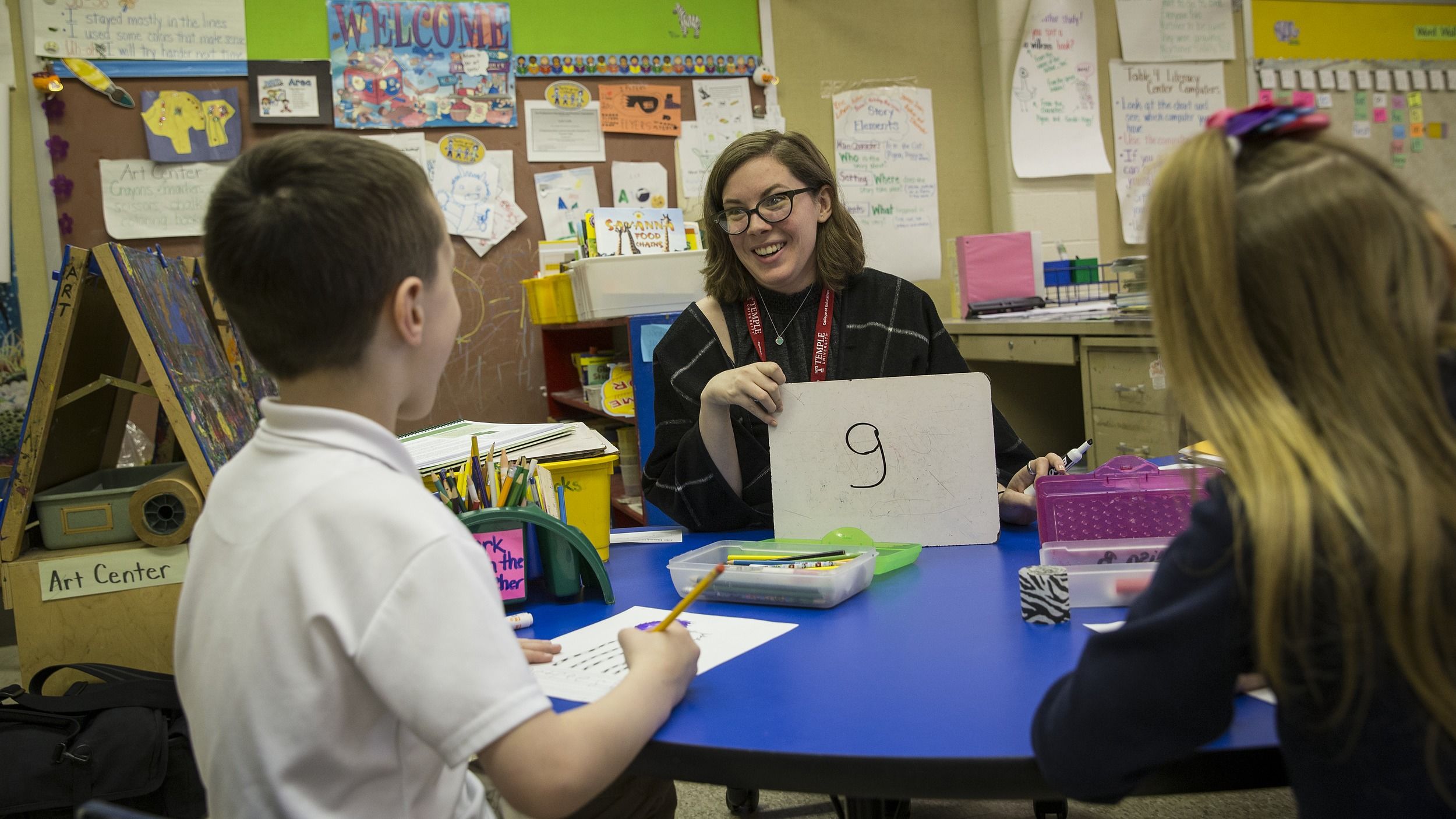 A student teacher sits at a desk with students in an elementary classroom.