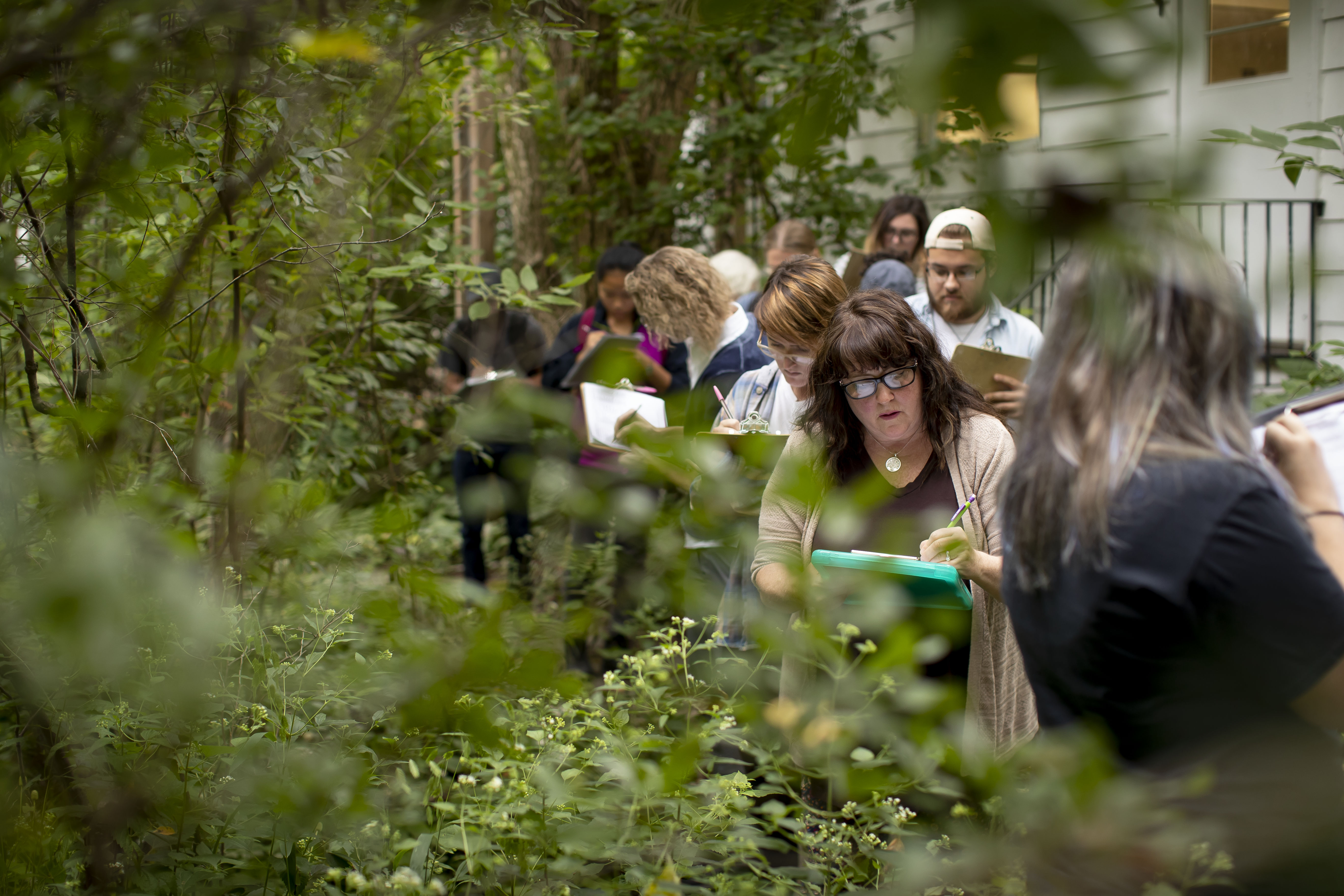 Students stand next to plants jotting down notes on clipboards.