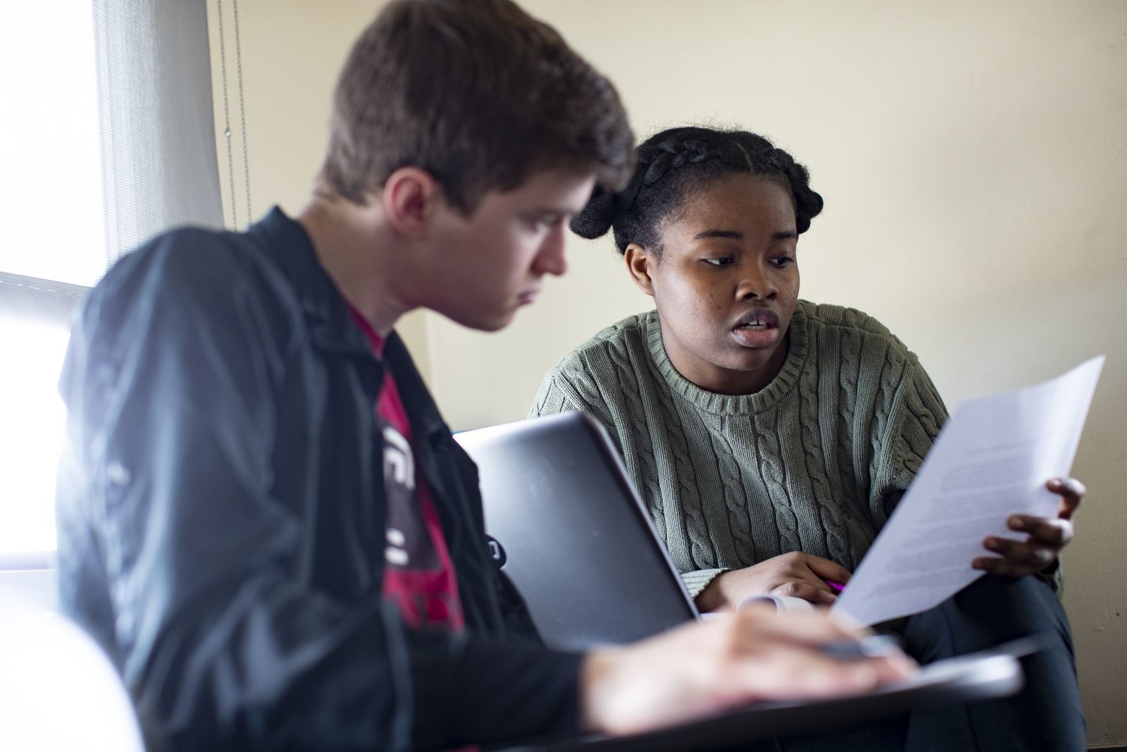 Two students look at a paper while sitting at desks in a classroom.
