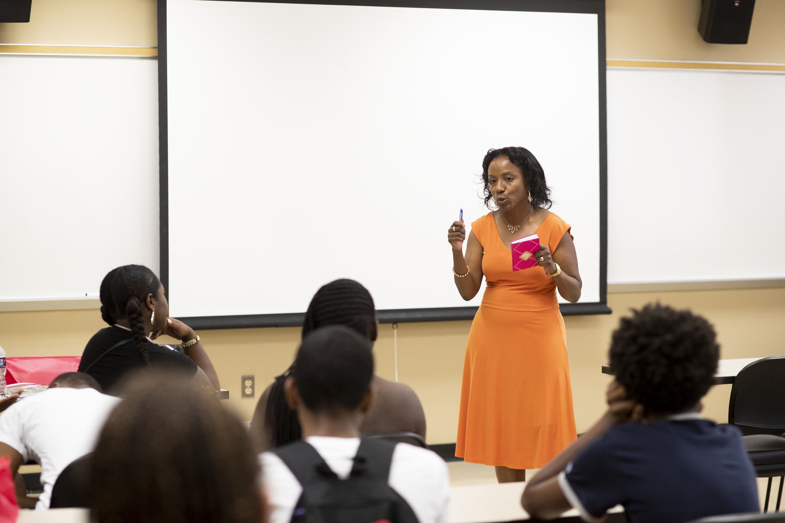 A professor in an orange dress lectures at the front of a classroom.