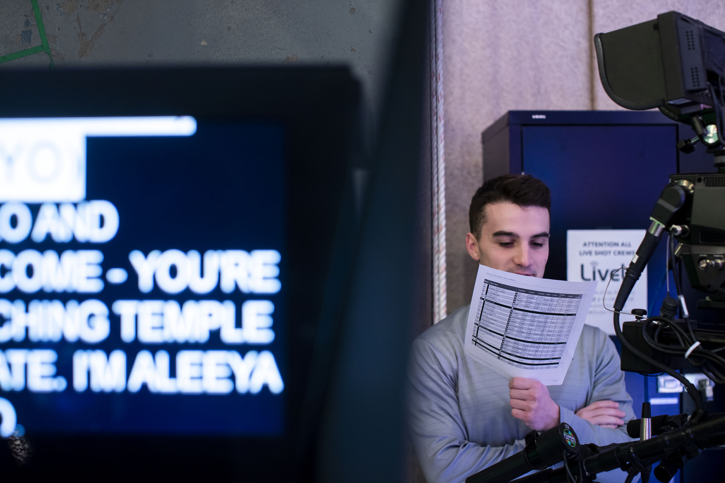 A student stands against the wall of a broadcasting studio reading a script while a monitor in front displays text.